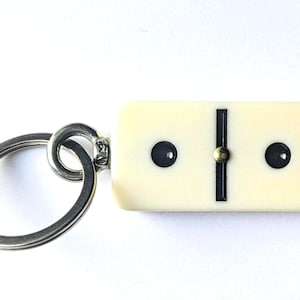 May include: A keychain featuring a rectangular, off-white domino tile with two black pips and a central black line. The domino is attached to a circular silver-toned metal split ring by a small metal connector loop, creating a functional accessory.
