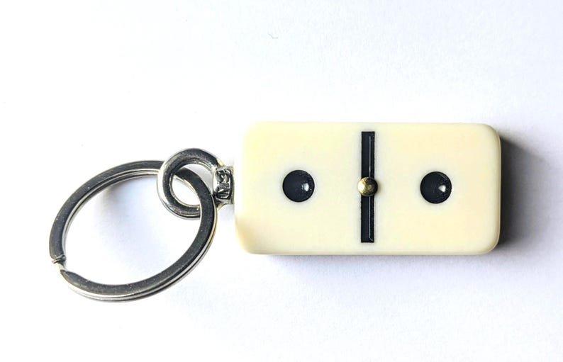 May include: A keychain featuring a rectangular, off-white domino tile with two black pips and a central black line. The domino is attached to a circular silver-toned metal split ring by a small metal connector loop, creating a functional accessory.
