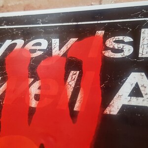 Op de afbeelding: Een close-up van een verweerd bord met de woorden "Coney Island" en "Stillwell Ave" in witte letters op een zwarte achtergrond. Een rode handafdruk bedekt een deel van het bord.