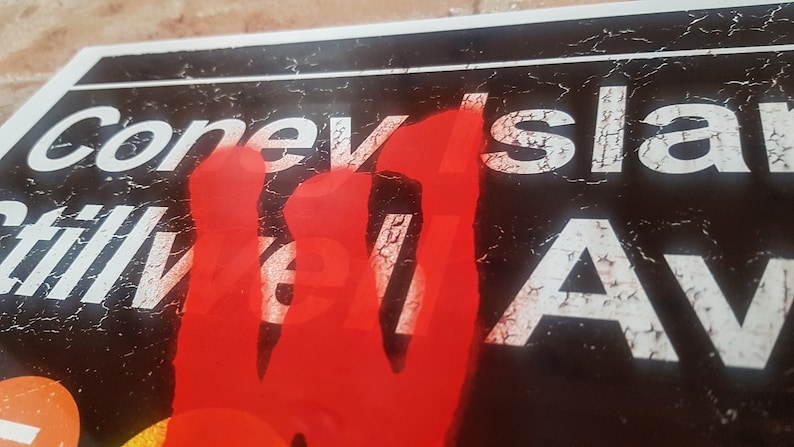 Op de afbeelding: Een close-up van een verweerd bord met de woorden "Coney Island" en "Stillwell Ave" in witte letters op een zwarte achtergrond. Een rode handafdruk bedekt een deel van het bord.