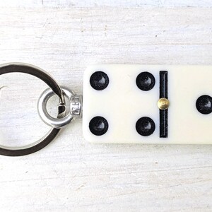 May include: A keychain featuring a rectangular off-white domino tile with five black pips and a central brass-colored pin. The domino is attached to a circular silver-toned metal split ring by a small metal connector loop.