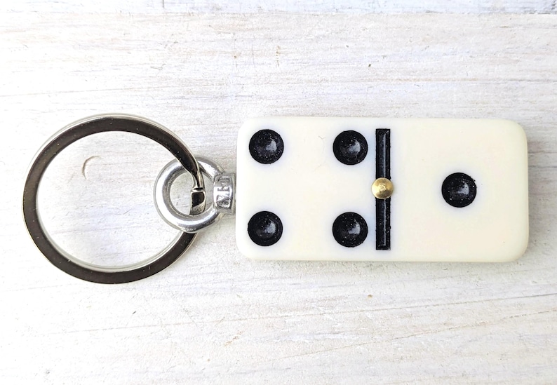 May include: A keychain featuring a rectangular off-white domino tile with five black pips and a central brass-colored pin. The domino is attached to a circular silver-toned metal split ring by a small metal connector loop.
