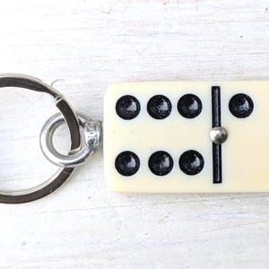May include: A keychain featuring a rectangular off-white domino tile with black pips. The domino displays a six on one side and a one on the other, separated by a black line and a small silver-toned metal rivet. A circular silver metal ring is attached.
