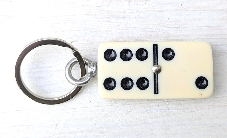 May include: A keychain featuring a rectangular off-white domino tile with black pips. The domino displays a six on one side and a one on the other, separated by a black line and a small silver-toned metal rivet. A circular silver metal ring is attached.