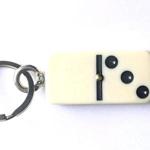 May include: A keychain featuring an off-white rectangular domino tile with three black circular pips on one side. The domino is attached to a circular silver-toned metal split ring by a small metal connector and a tiny gold-colored rivet.