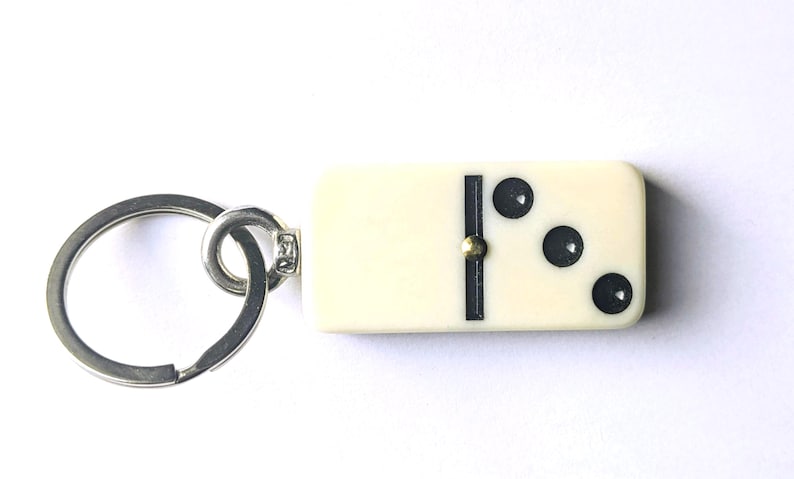 May include: A keychain featuring an off-white rectangular domino tile with three black circular pips on one side. The domino is attached to a circular silver-toned metal split ring by a small metal connector and a tiny gold-colored rivet.