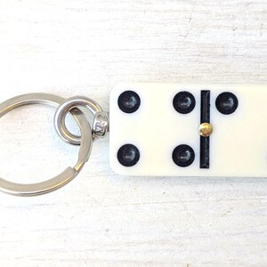 May include: A keychain featuring a rectangular off-white domino tile with black pips and a central gold-toned metal pin. The domino displays a total of seven black dots arranged in a specific pattern, attached to a circular silver-toned metal ring.