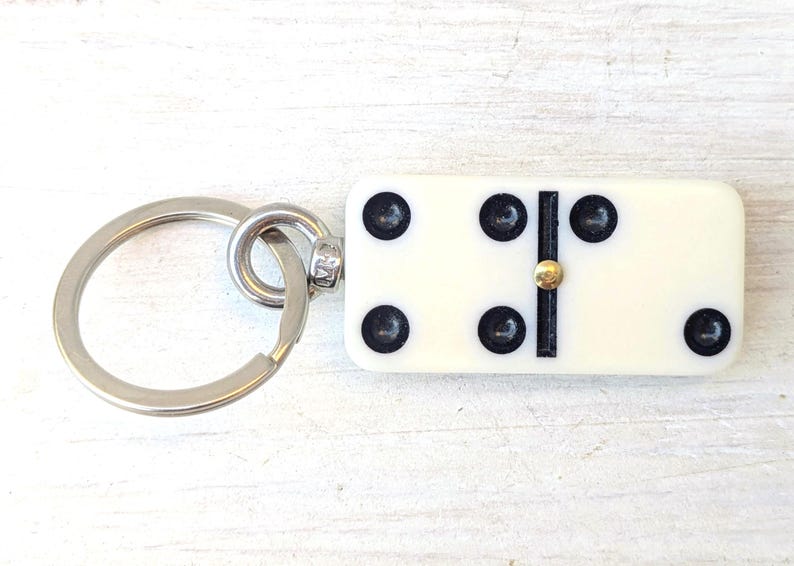 May include: A keychain featuring a rectangular off-white domino tile with black pips and a central gold-toned metal pin. The domino displays a total of seven black dots arranged in a specific pattern, attached to a circular silver-toned metal ring.