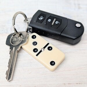 May include: A metal keyring holding a silver Yale key, a black car key fob with three buttons, and a cream-colored domino tile with black pips. The domino features a central metal pin and displays a five-pip pattern on its visible surface.