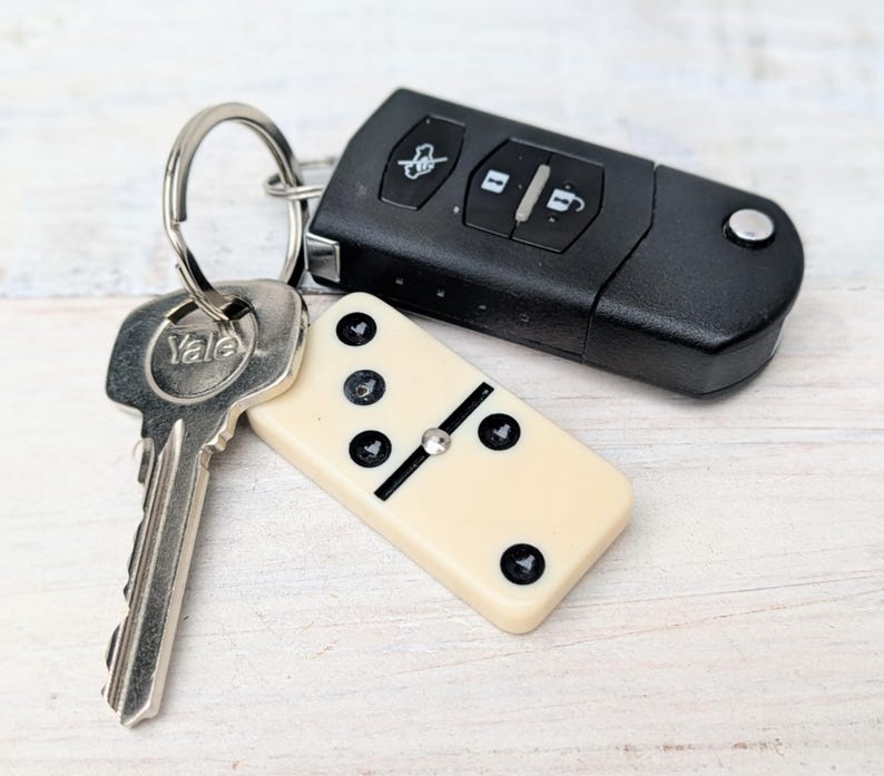 May include: A metal keyring holding a silver Yale key, a black car key fob with three buttons, and a cream-colored domino tile with black pips. The domino features a central metal pin and displays a five-pip pattern on its visible surface.