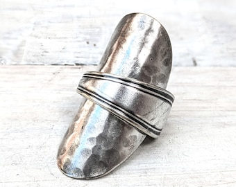 Könnte beinhalten: Ein breiter silberfarbener Ring, der aus einem recycelten Metalllöffelgriff gefertigt wurde. Der Ring weist eine gehämmerte, texturierte Oberfläche am länglichen Schaft und ein glattes, erhabenes Banddetail in der Mitte auf, mit metallischem Glanz.