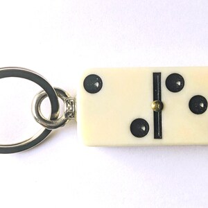 May include: A keychain featuring an off-white rectangular domino tile with black pips and a central black divider line. The domino displays a two-pip side and a three-pip side, connected to a circular silver-toned metal split ring by a small metal loop.