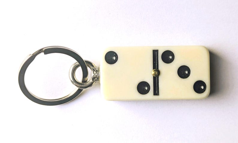 May include: A keychain featuring an off-white rectangular domino tile with black pips and a central black divider line. The domino displays a two-pip side and a three-pip side, connected to a circular silver-toned metal split ring by a small metal loop.