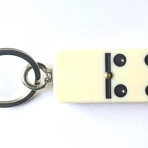 May include: A keychain featuring a rectangular off-white domino tile with four black pips and a central black divider line. The domino is attached to a circular silver-toned metal split ring by a small metal jump ring and a connector pin.
