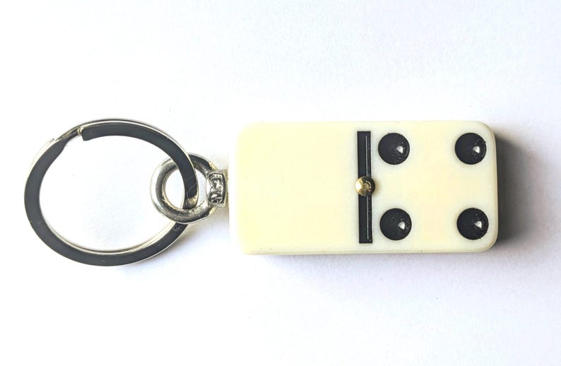 May include: A keychain featuring a rectangular off-white domino tile with four black pips and a central black divider line. The domino is attached to a circular silver-toned metal split ring by a small metal jump ring and a connector pin.