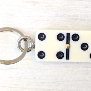 May include: A keychain featuring a rectangular off-white domino tile with black pips and a central gold-colored metal pin. The domino is attached to a circular silver-toned metal split ring by a small metal jump ring and a short connector piece.
