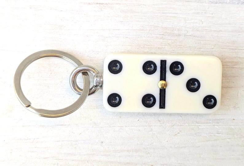 May include: A keychain featuring a rectangular off-white domino tile with black pips and a central gold-colored metal pin. The domino is attached to a circular silver-toned metal split ring by a small metal jump ring and a short connector piece.