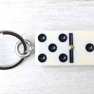 May include: A keychain featuring a rectangular off-white domino tile with black pips and a central gold-colored metal pin. The domino is attached to a circular silver-toned metal split ring by a small metal jump ring and a silver-colored connector.
