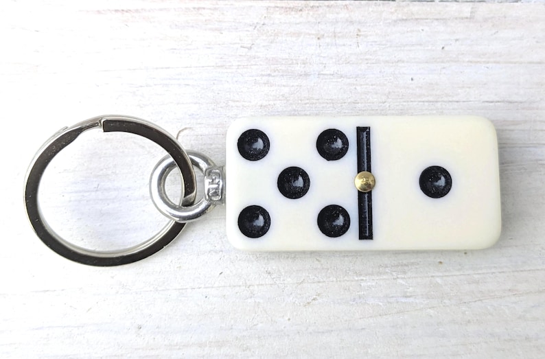 May include: A keychain featuring a rectangular off-white domino tile with black pips and a central gold-colored metal pin. The domino is attached to a circular silver-toned metal split ring by a small metal jump ring and a silver-colored connector.