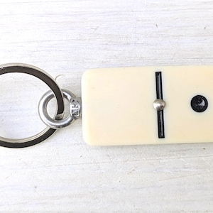 May include: A keychain featuring a rectangular, off-white domino tile with a single black dot and a central black line divider. The domino is attached to a circular silver-toned metal split ring by a small metal eyelet and a jump ring connector.
