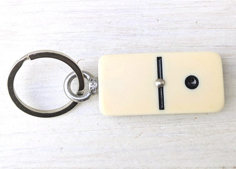 May include: A keychain featuring a rectangular, off-white domino tile with a single black dot and a central black line divider. The domino is attached to a circular silver-toned metal split ring by a small metal eyelet and a jump ring connector.