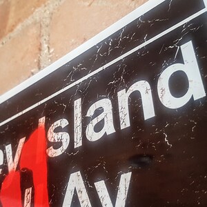 Op de afbeelding: Een zwart-wit bord met de woorden "Coney Island Ave" in witte letters. Het bord is verweerd en heeft een rode vlamgrafiek.