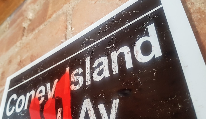Op de afbeelding: Een zwart-wit bord met de woorden "Coney Island Ave" in witte letters. Het bord is verweerd en heeft een rode vlamgrafiek.