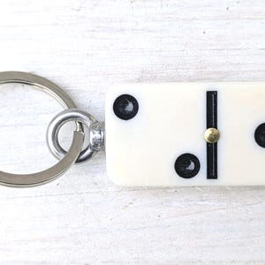 May include: A keychain featuring a rectangular off-white domino tile with three black pips and a central black line. The domino is attached to a circular silver-toned metal split ring by a small metal eyelet and a tiny brass-colored rivet.