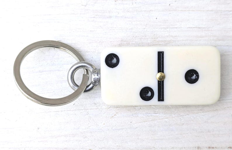 May include: A keychain featuring a rectangular off-white domino tile with three black pips and a central black line. The domino is attached to a circular silver-toned metal split ring by a small metal eyelet and a tiny brass-colored rivet.