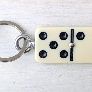May include: A keychain featuring a rectangular off-white domino tile with black pips. The domino displays a five-pip pattern on the left side and a single pip on the right side, separated by a black line. A silver-toned metal ring is attached.