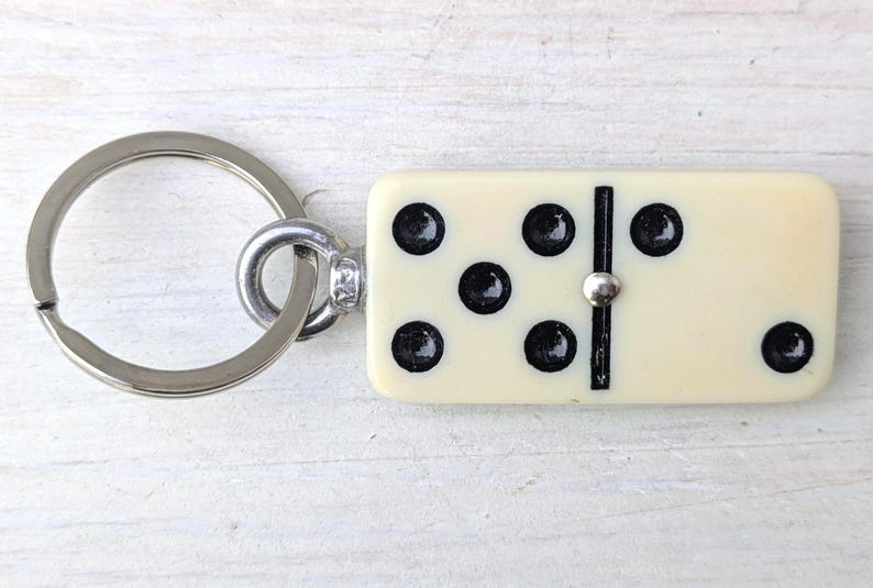 May include: A keychain featuring a rectangular off-white domino tile with black pips. The domino displays a five-pip pattern on the left side and a single pip on the right side, separated by a black line. A silver-toned metal ring is attached.