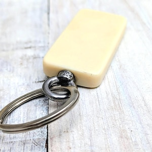 May include: A rectangular, off-white, smooth-surfaced keychain tag attached to a circular, dark metallic split ring. The tag features a flat, solid-colored face and is connected to the ring by a small, cylindrical metal hardware component.