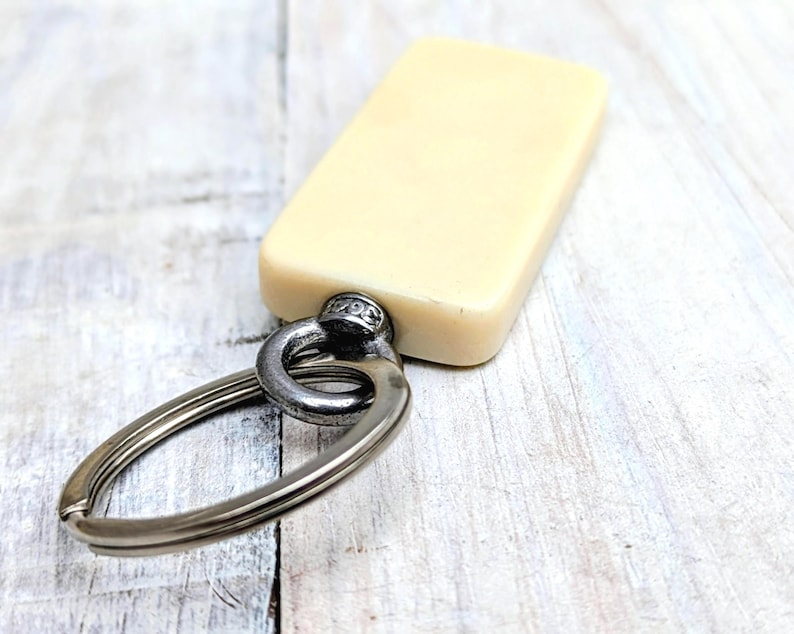 May include: A rectangular, off-white, smooth-surfaced keychain tag attached to a circular, dark metallic split ring. The tag features a flat, solid-colored face and is connected to the ring by a small, cylindrical metal hardware component.