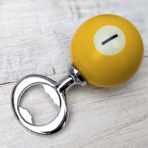 Może przedstawiać: Otwieracz do butelek z ż&oacute;łtą kulą bilardową, z białym okręgiem i czarnym paskiem. Otwieracz ma błyszczący, srebrny metalowy uchwyt.