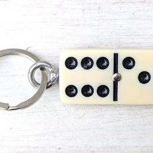 May include: A keychain featuring a rectangular off-white domino tile with black pips. The domino displays a six on the left side and a three on the right side, separated by a black line and a small silver-colored metal pin. A silver metal ring is attached.