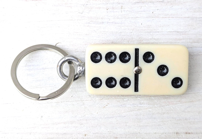 May include: A keychain featuring a rectangular off-white domino tile with black pips. The domino displays a six on the left side and a three on the right side, separated by a black line and a small silver-colored metal pin. A silver metal ring is attached.