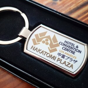 May include: A silver key chain with a white rectangular tag. The tag has a gold logo with three dots and the text "Hotel & Convention Centre Los Angeles Nakatomi Plaza 中音プラザ".