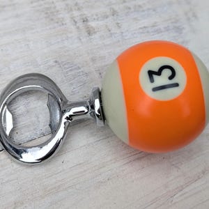 Może przedstawiać: Chromowany otwieracz do butelek z motywem kuli bilardowej z numerem 13. Kula jest pomarańczowo-kremowa z czarnym numerem 13. Otwieracz wykonany jest z błyszczącego chromu.
