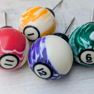 May include: Five cabinet knobs designed as billiard balls. Each knob has a different colour scheme, including red, orange, purple, green, and white, with black numbering. The knobs have a silver metal stem.