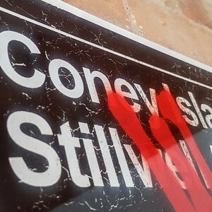 Op de afbeelding: Een zwart-wit bord met de woorden "Coney Island Stillwell Av" in witte letters. Het bord heeft een rode spuitverfmarkering over het woord "Stillwell".