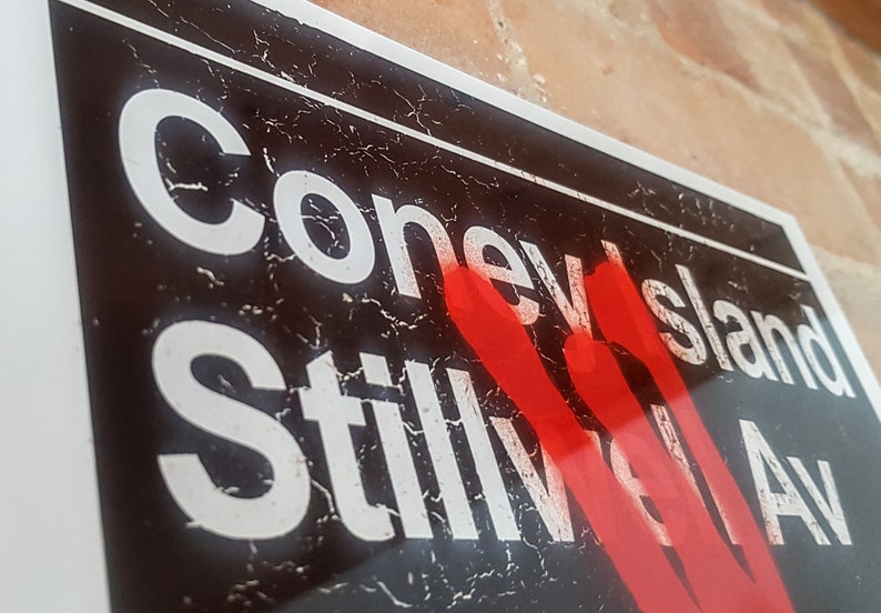 Op de afbeelding: Een zwart-wit bord met de woorden "Coney Island Stillwell Av" in witte letters. Het bord heeft een rode spuitverfmarkering over het woord "Stillwell".