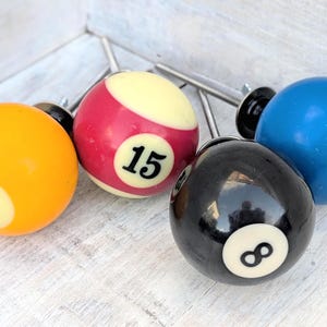 May include: Four spherical cabinet knobs designed to resemble billiard balls. The set includes a yellow ball with the number 1, a red and white ball with the number 15, a black ball with the number 8, and a blue ball with the number 2, each with a metal bolt.