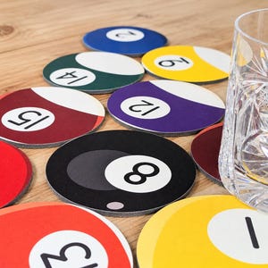 May include: A collection of round drink coasters designed to look like colorful billiard balls, each featuring a specific number and a white circular accent. The set includes red, yellow, blue, green, purple, and black coasters alongside a glass.