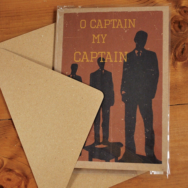 May include: A greeting card with the text "O Captain My Captain" in gold lettering, featuring black silhouettes of men on a reddish-brown background. The card is inside a clear plastic sleeve, with a kraft paper envelope.