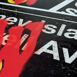 Op de afbeelding: Een zwart-wit bord met de woorden "Cornell Island Av" in witte letters. Het bord is verweerd en heeft een rode handafdruk aan de linkerkant.