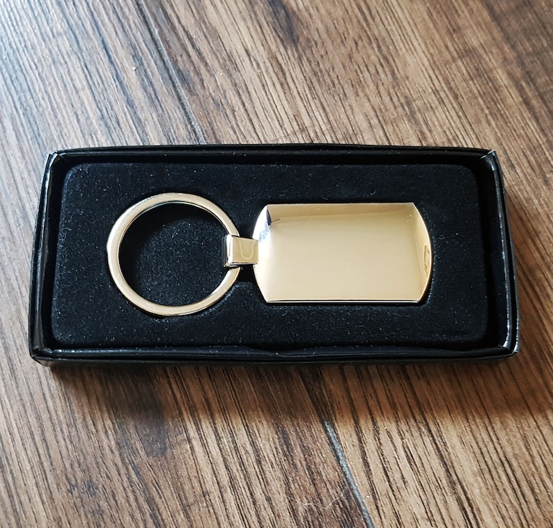 May include: A silver key chain with a blank rectangular tag. The keychain is in a black velvet box.