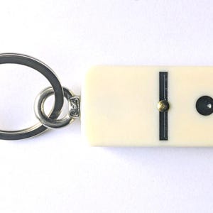 May include: A keychain featuring a rectangular, off-white domino tile with a single black dot and a vertical black line. The domino is attached to a circular silver-toned metal split ring by a small metal jump ring and a short, flat metal connector.