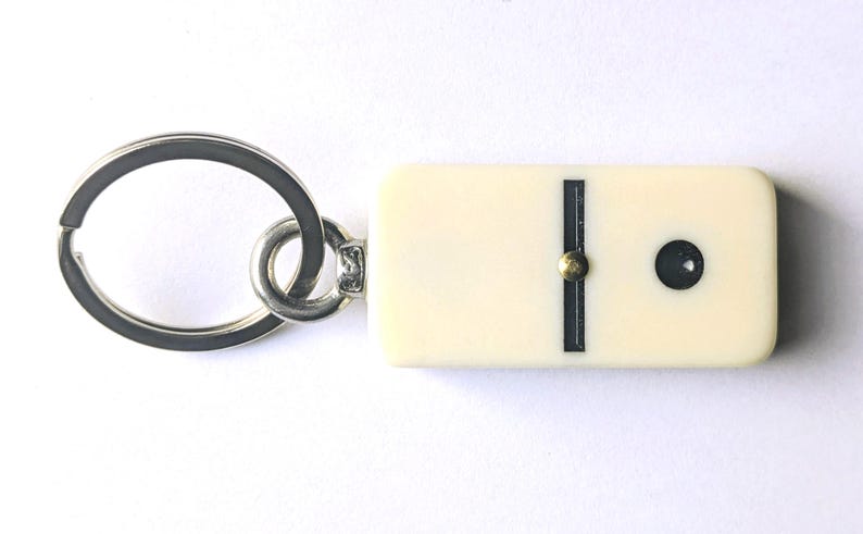 May include: A keychain featuring a rectangular, off-white domino tile with a single black dot and a vertical black line. The domino is attached to a circular silver-toned metal split ring by a small metal jump ring and a short, flat metal connector.