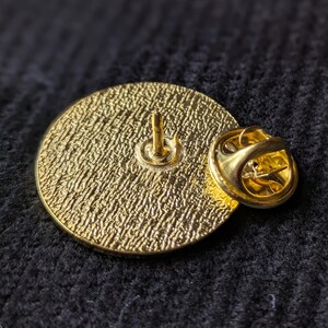 May include: The back of a gold-coloured pin with a textured surface and a pin back.