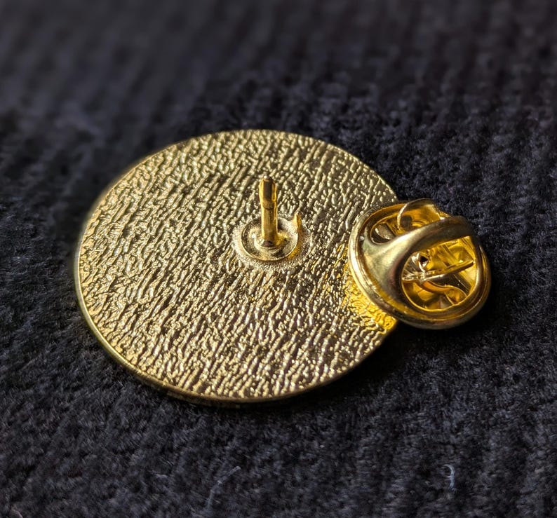 May include: The back of a gold-coloured pin with a textured surface and a pin back.